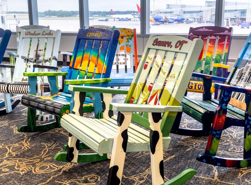 Colourful hand-painted Adirondack rocking chairs representing Connecticut towns including Somers, Coventry, and Windsor Locks, displayed in an airport terminal.