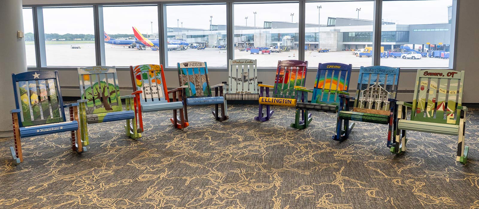 A collection of vibrantly painted rocking chairs representing Connecticut towns — including Ellington, Windsor, Glastonbury, and Somers — arranged in a circle at Bradley International Airport, with planes visible through the terminal windows.