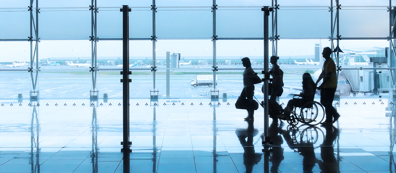 Four people, including one in a wheelchair, move through an airport terminal with large windows showing the tarmac and planes outside.