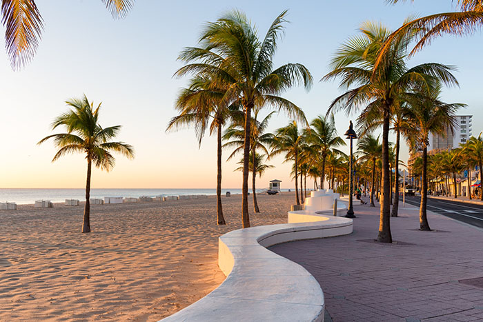 Palm trees line a curving white promenade along the sandy beach in Fort Lauderdale at sunrise.