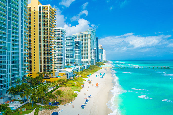 High-rise beachfront towers overlook turquoise waters and a sandy shore dotted with beachgoers in Miami.