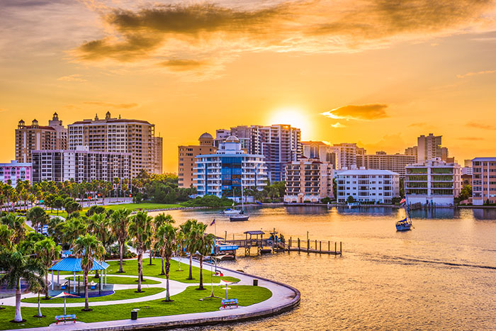 A golden sunset silhouettes Sarasota's waterfront skyline behind a landscaped park and marina.