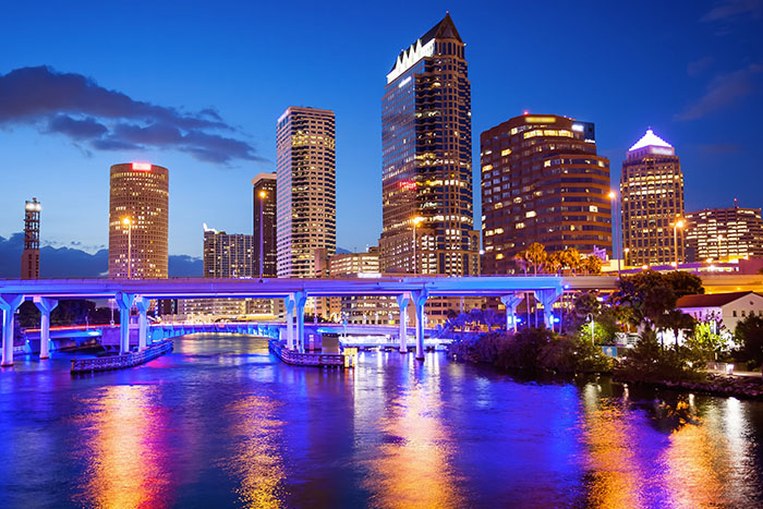 Tampa's illuminated downtown skyline reflects on the river at dusk, framed by a lit pedestrian bridge.