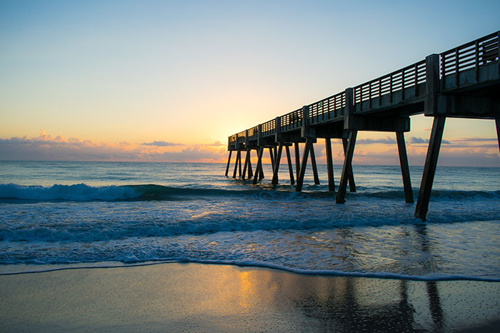 A wooden fishing pier stretches over gentle waves as the sun rises over the horizon in Vero Beach.