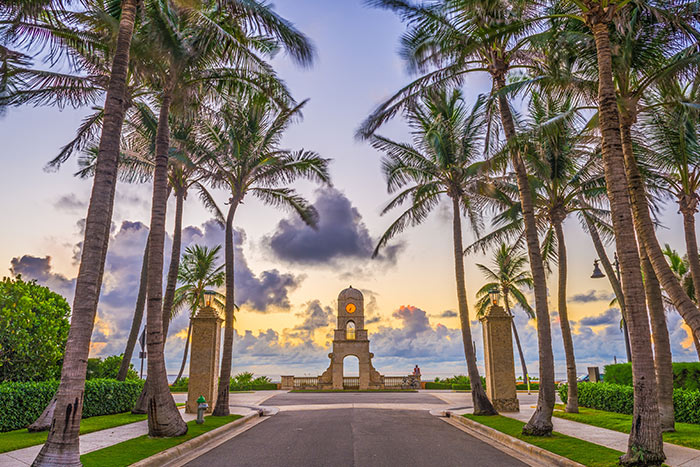 A palm-lined pathway leads to the iconic Worth Avenue clock tower archway in West Palm Beach at sunset.
